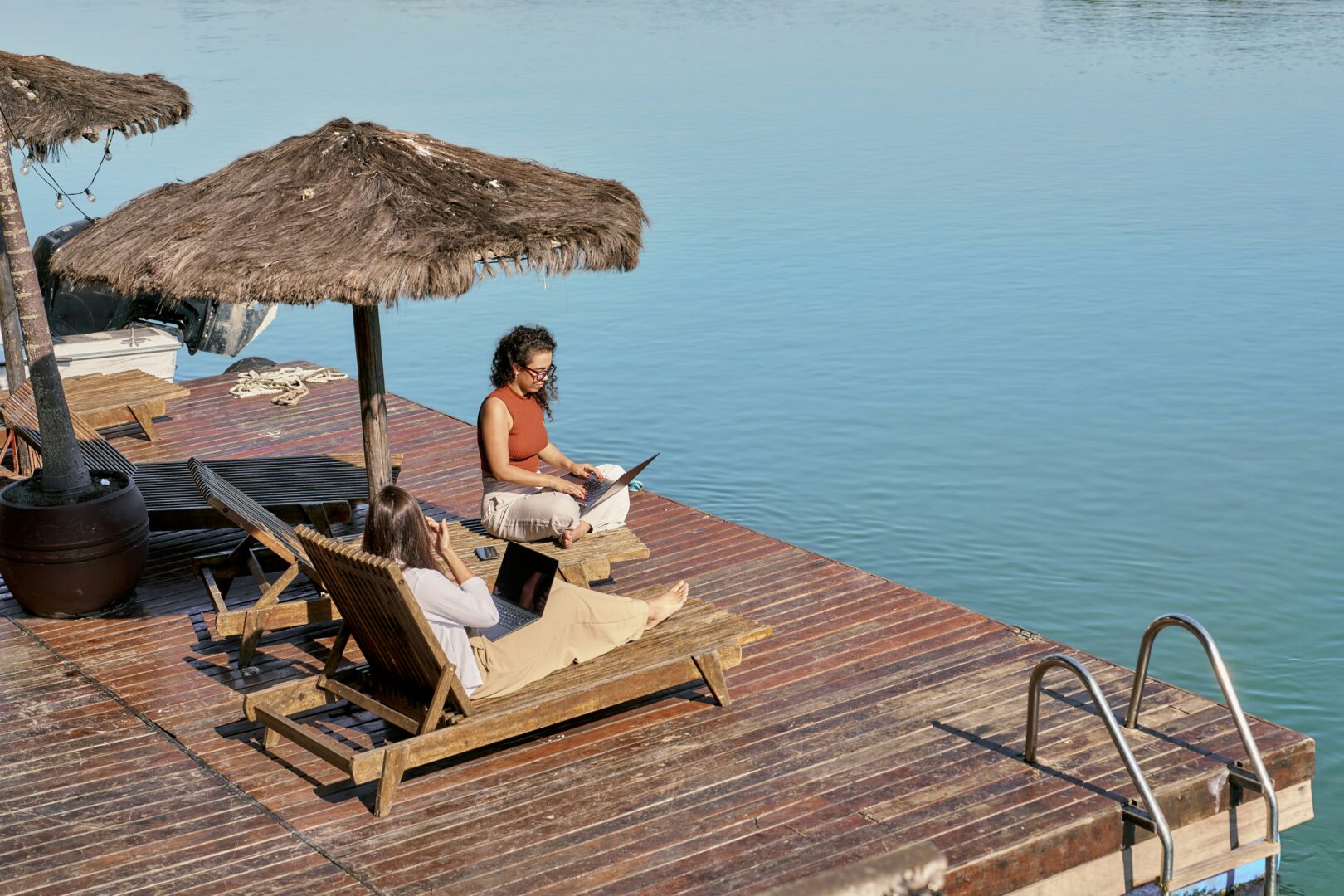 Two women sitting on a deck near a lake working on their laptops