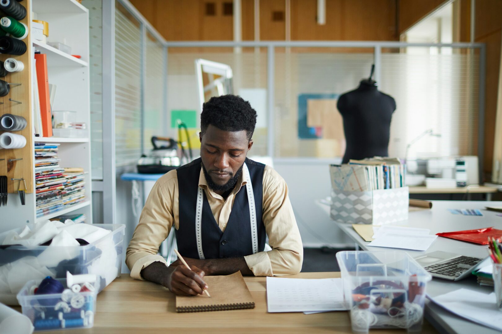 A man sitting at his desk drawing in a book with a measuring tape around is neck