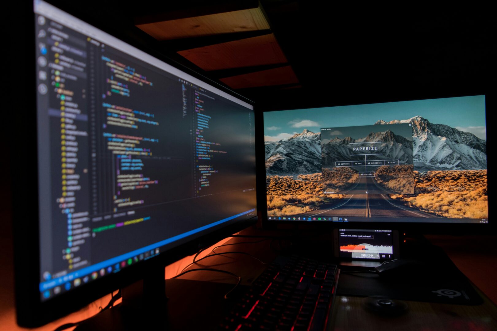 A computer screen with coding on it and another one with a background of a mountain in a dark room